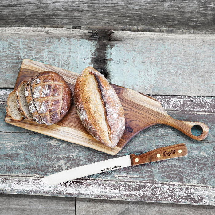 Top view of teak cutting board with loafs and a bread knife displayed on gray, wooden surface
