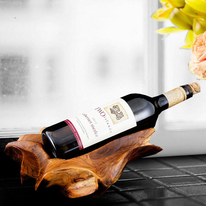 A teak wine bottle holder with an irregular shape, holding a wine bottle, placed on a table with a window in the background.