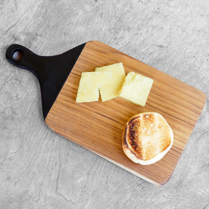Top view of a teak cutting board with black handle with cheese and a piece of bread on a gray surface