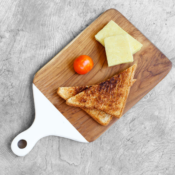 Top view of squared shaped teak cutting board/serving platter with white handle with toast, cheese, and tomatoes on a light gray wooden surface