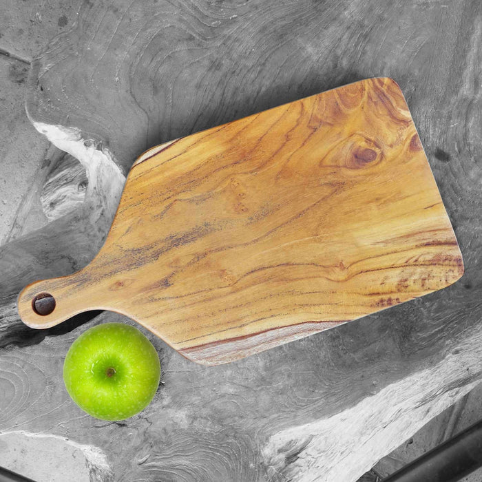 Top view of squared teak cutting board with handle next to a green apples on a light gray marble surface