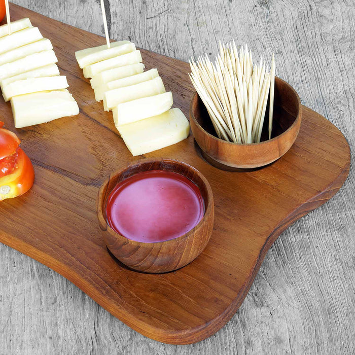 Teak serving plater with 2 teak bowls with red liquid and toothpicks inside the bowls next to rows of cheese and tomatoes displayed on a wooden surface
