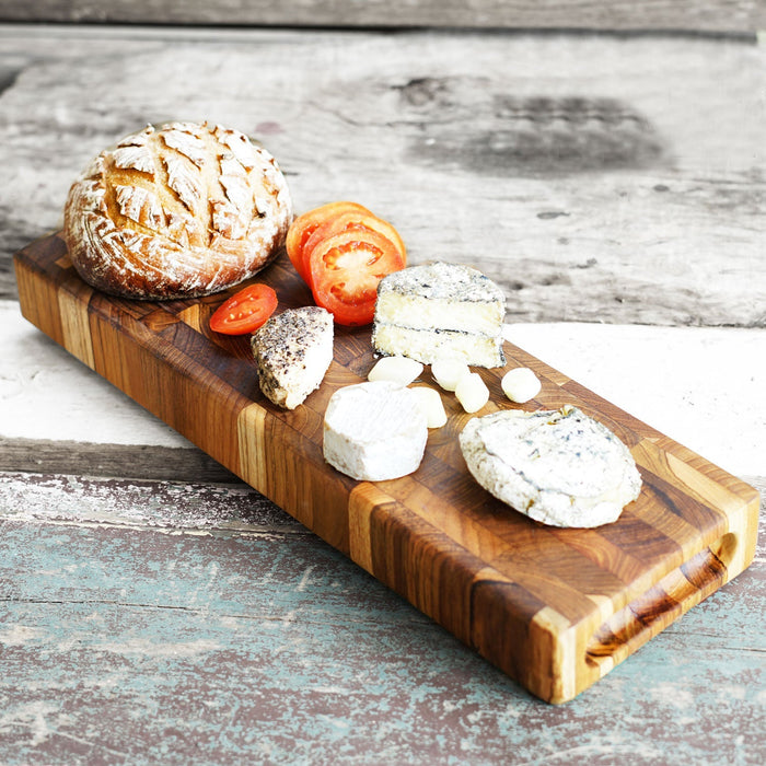 Rectangular end grain teak cutting board with bread, sliced tomatoes, and cheese displayed on a wooden surface