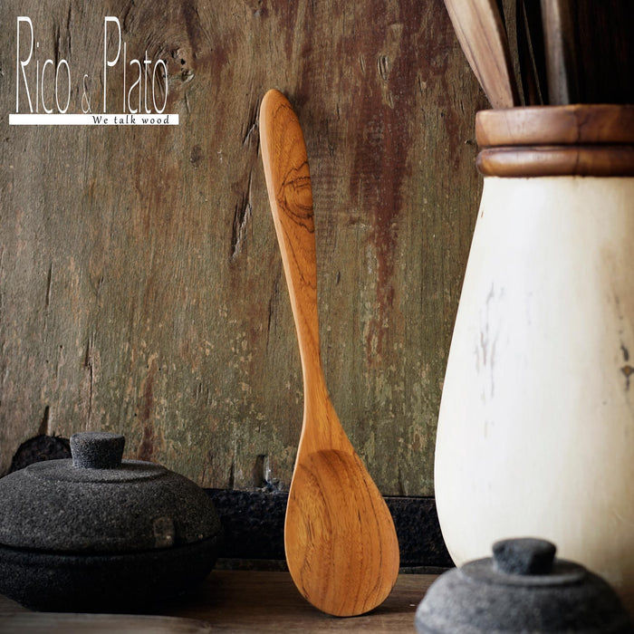 A handcrafted teak wood spoon with a natural finish, leaning against a rustic wooden surface, with a white ceramic container and a black pot in the background.