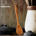 A handcrafted teak wood spoon with a natural finish, leaning against a rustic wooden surface, with a white ceramic container and a black pot in the background.