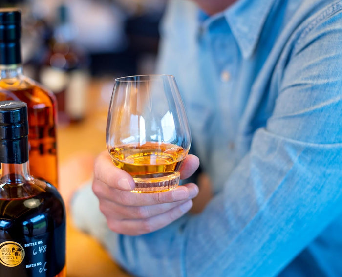 Person holding a glass of bourbon with bottles in the background