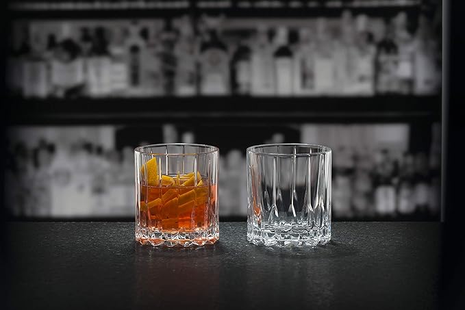 Two Riedel glassware neat glass one with brown liquid inside and one empty on a black countertop and bar setting in the background