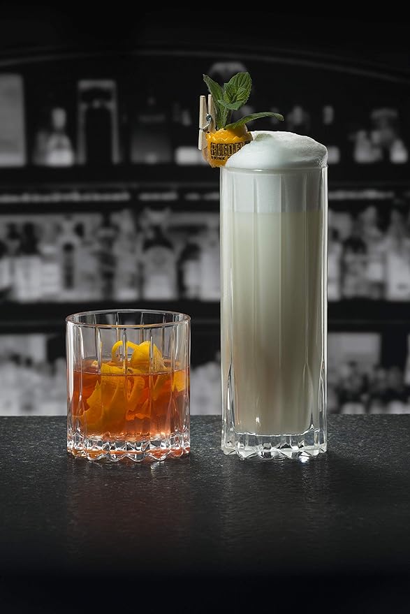 Two Riedel glassware neat glasses with creamy drink inside a tall neat glass and brown liquid inside a short neat glass on a dark countertop and bar background