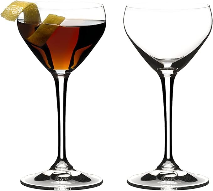 Set of two of Riedel drink specific glassware Nick & Nora cocktail glasses one with brown liquid and lemon peel inside and other is empty on a white background