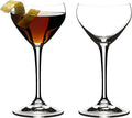 Set of two of Riedel drink specific glassware Nick & Nora cocktail glasses one with brown liquid and lemon peel inside and other is empty on a white background