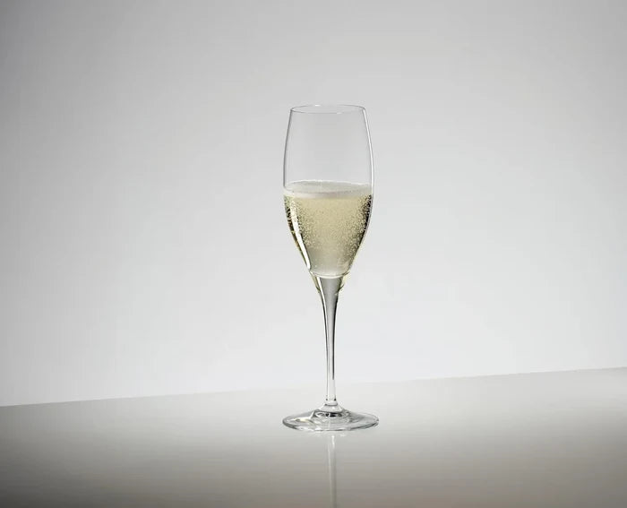 A clear champagne flute glass partially filled with pale golden bubbly beverage, centered on a light gray studio background with a reflective surface below.