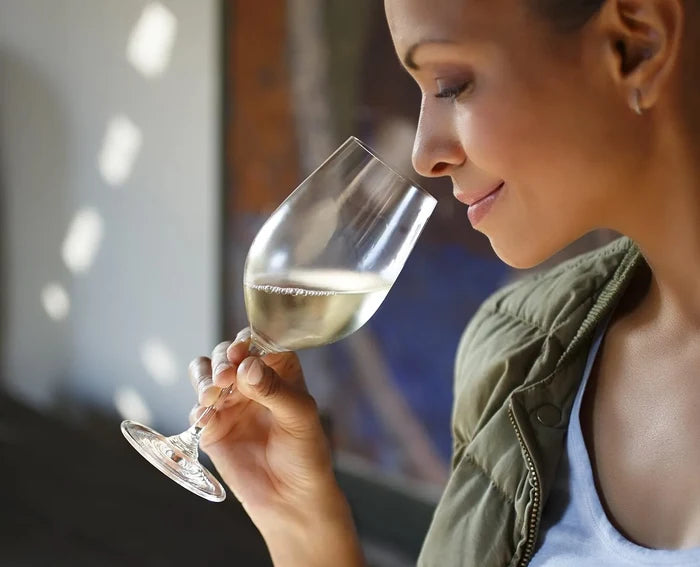 A woman in profile holding a wine glass with white wine, wearing an olive green jacket over a white top, in a softly lit indoor setting.