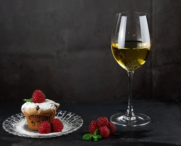 A stemmed wine glass filled with white wine stands on a dark surface beside a small dessert cupcake on a clear plate, with raspberries and mint leaves scattered nearby against a dark textured background.