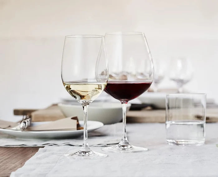 Two clear wine glasses on a table, one filled with white wine and one with red wine, alongside a clear drinking glass in a softly lit dining setting.