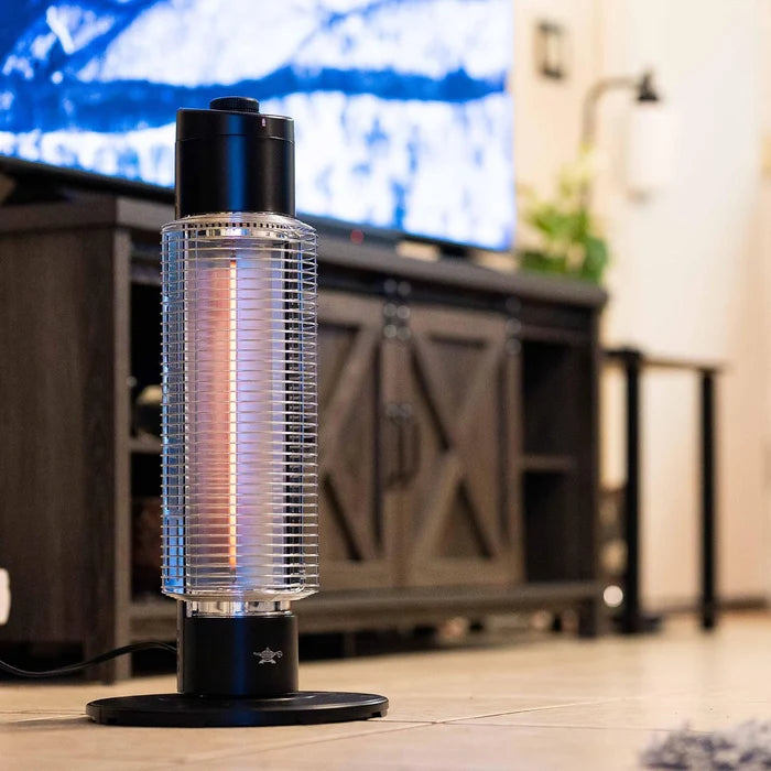 Tall, black cylindrical heater on a wooden floor with a blurred TV and furniture in the background