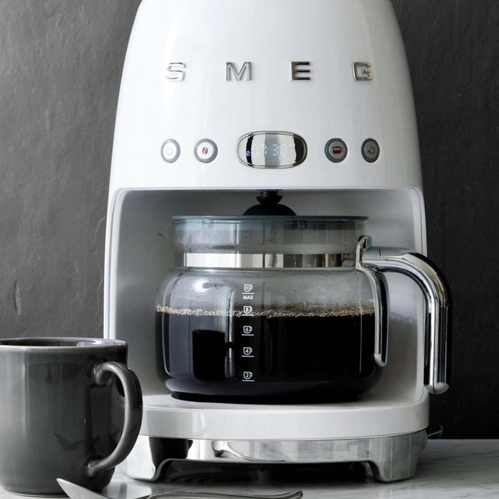 White Smeg drip coffee machine with filled coffee glass carafe on a marble surface and gray background