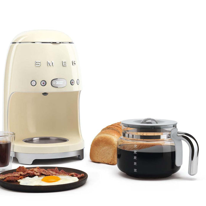 Cream Smeg 50s retro line drip coffee machine next to breakfast and a coffee filled glass carafe displayed on a white background