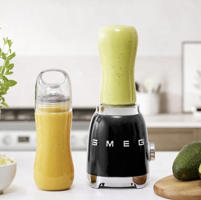 Black Smeg retro line personal blender next to a clear tumbler both with smoothies inside on a white countertop