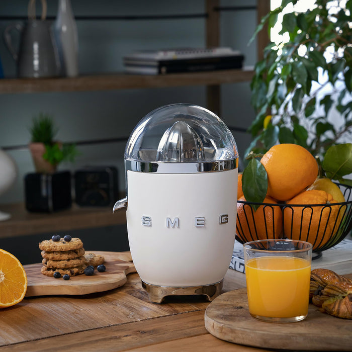 White Smeg juicer on a wooden table with oranges and juice.