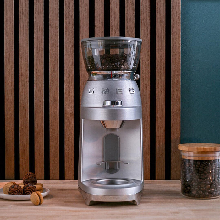 Stainless steel Smeg retro style electric espresso coffee grinder next to a jar of coffee beans and snacks on a wooden surface