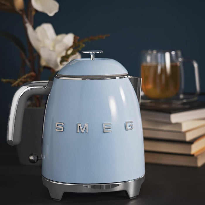 Light blue Smeg kettle on a dark surface with books and a glass of tea in the background.