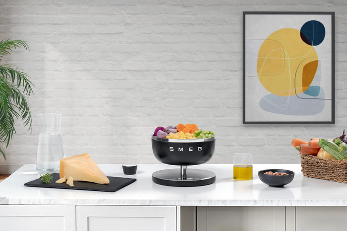 Matte black Smeg contemporary kitchen scale weighing fruit and veggies next to cheese, fruit, veggies, and drinks on a white countertop