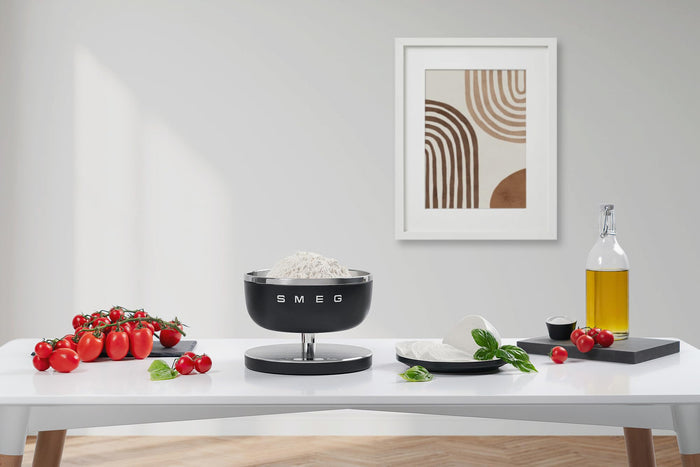 Matte black Smeg contemporary kitchen scale weighing flour next to oil, tomatoes, and cheese on a white tabletop