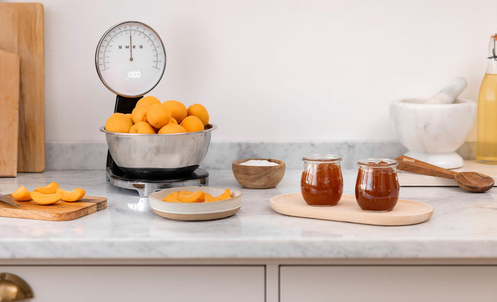 Black, silver Smeg retro style analog digital kitchen scale with fruit inside the silver bowl of the scale with food and jam around the scale on a marble counter in a kitchen setting