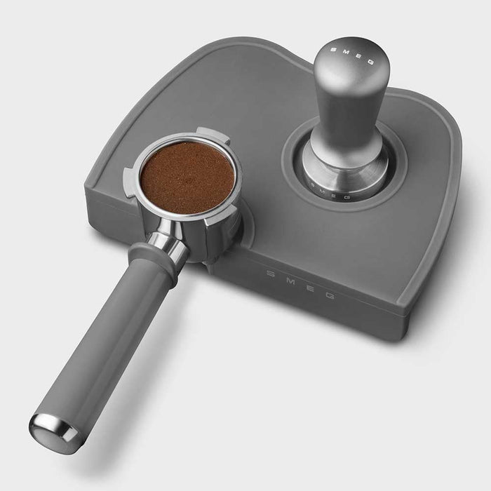 Grey, stainless steel Smeg tamper and portafilter with ground coffee grinds inside on a white background