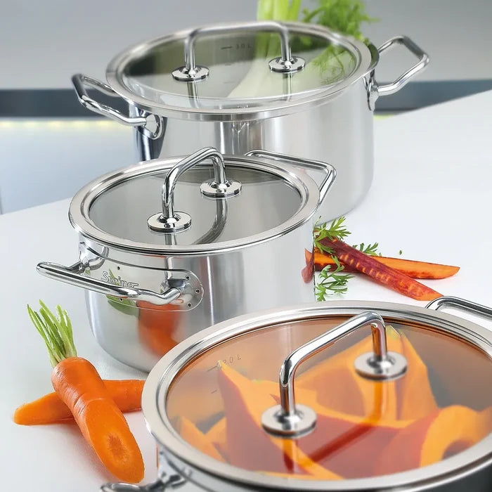 Three stainless steel stock pots with glass lids are arranged on a white countertop with fresh carrots and herbs as props.