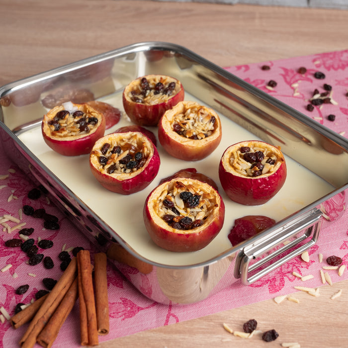 Stuffed apples in a baking dish on a pink floral tablecloth with cinnamon sticks and raisins.