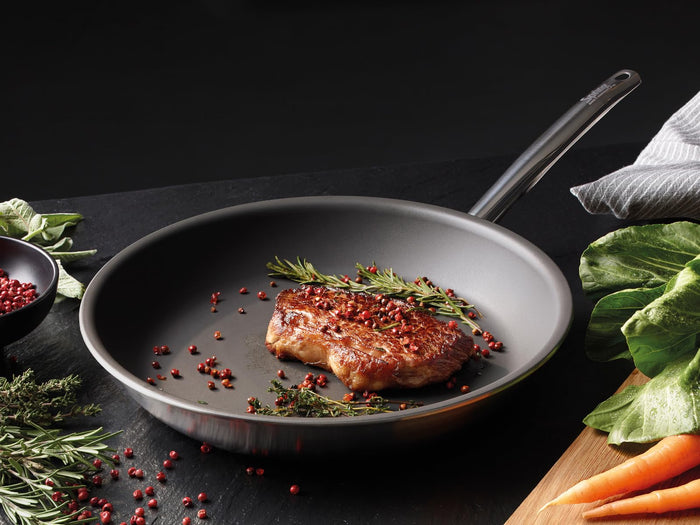 Pan with seared meat and herbs on a dark surface with vegetables and spices.