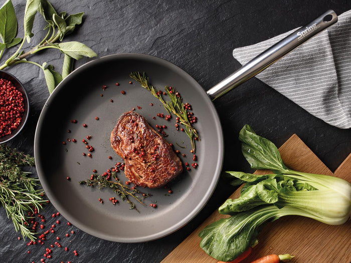 Frying pan with meat and herbs on a dark surface with vegetables around