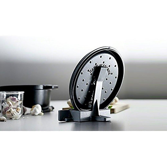 Staub Cast Iron Lid Holder in black holding a lid next to cooking materials and food displayed on a grey surface and background