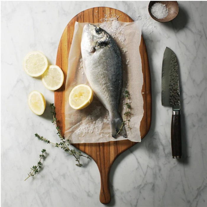 Teakhaus large canoe paddle cutting board with fish, salt, and lemon next to a knife displayed on a marble surface