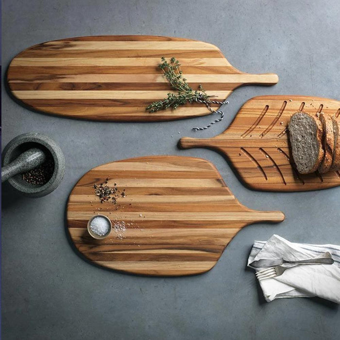 3 types of Teakhaus large canoe paddle cutting board displayed with salt, herbs, and bread on a grey surface