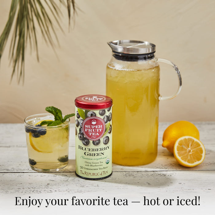 Blueberry Green tea next to glass of tea, jar of green tea, and lemons displayed on a white wooden surface