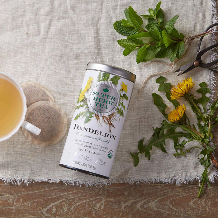Top view of Super Herb Tea Dandelion next to a mug of tea, 2 tea bags, and herbs on a white cloth on a table