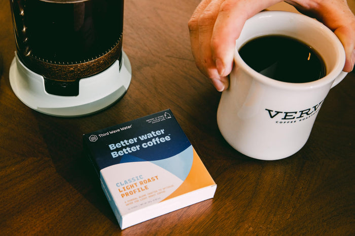 Hand holding a white mug with coffee, next to a box of 'Third Wave Water' and French press on a wooden table.