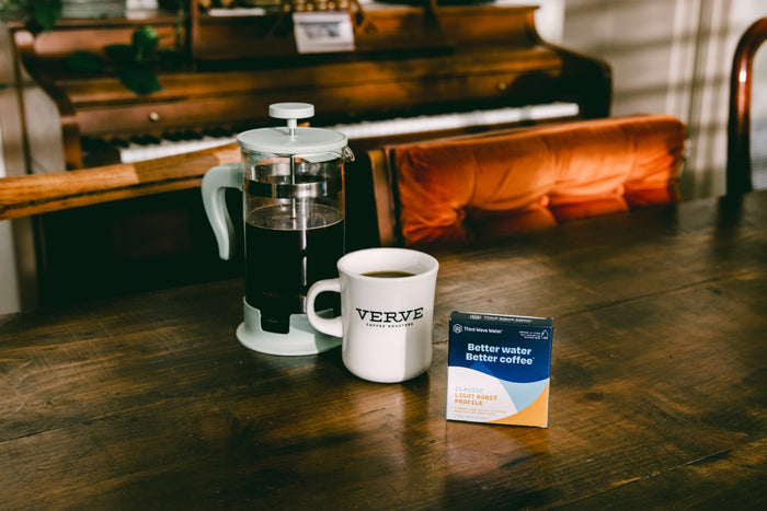 French press with coffee, a cup, and Third Wave Water Classic Light Roast Profile box on a wooden table in front of a piano