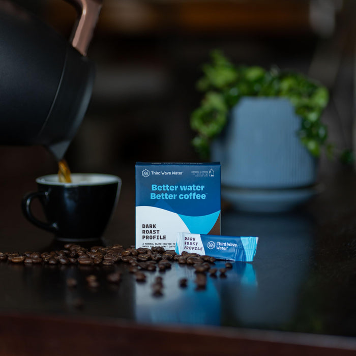 Box of Third Wave Water with coffee beans and a kettle pouring into a cup of coffee on a table.