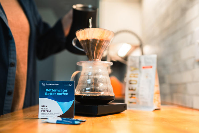 Coffee brewing setup with a person holding a coffee pot, a filter, and a box of Third Wave Water Dark Roast Profile on a wooden surface.