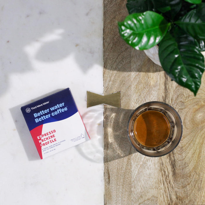 Box of Third Wave Water espresso package on a marble surface with a plant and cup of liquid on a wooden surface in the background