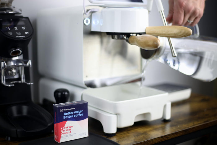 Espresso machine with a hand holding a clear pitcher, next to a box of Third Wave Water Espresso Machine Profile on a wooden surface.