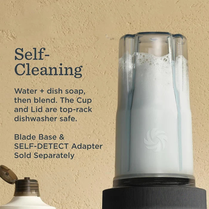 A close-up of a white and black cup-and-lid dishwasher safe blender base with a clear cup filled with soapy liquid, set against a textured beige background with text explaining the self-cleaning feature.