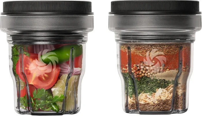Two clear plastic blender cups with black lids, shown side by side on a white background. The left cup contains fresh vegetables including tomato, green pepper, and herbs, while the right cup contains dry spices and ingredients layered vertically.