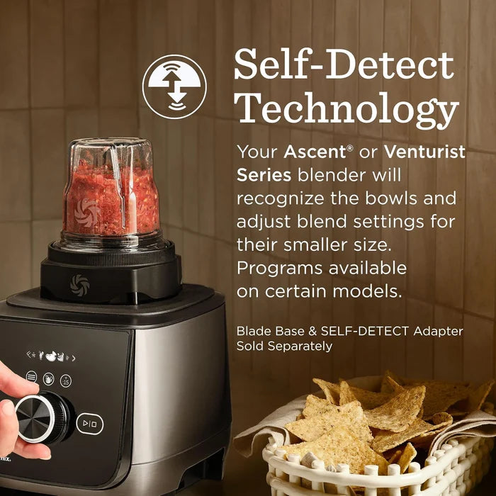 A countertop blender with a clear cup containing red food, shown beside marketing text about self-detect technology. A hand is turning the blender dial, and a basket of tortilla chips sits nearby against a brown tiled background.