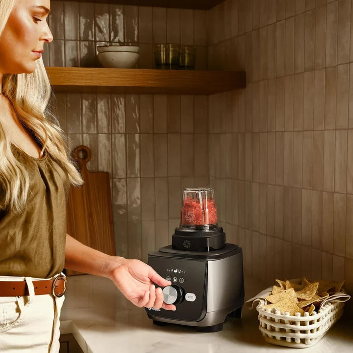A woman in an olive green top and white pants stands at a kitchen counter, reaching for the dial of a black countertop blender with a clear cup filled with red fruit. The kitchen has beige tiled walls, wooden shelves, and a basket of tortilla chips on the counter.