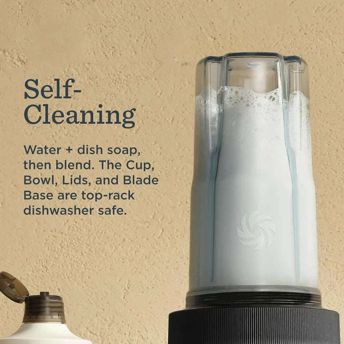 A countertop blender jar filled with white liquid, shown against a warm beige textured wall with text reading 'Self-Cleaning' and smaller cleaning instructions. Part of a dish soap bottle is visible at lower left.