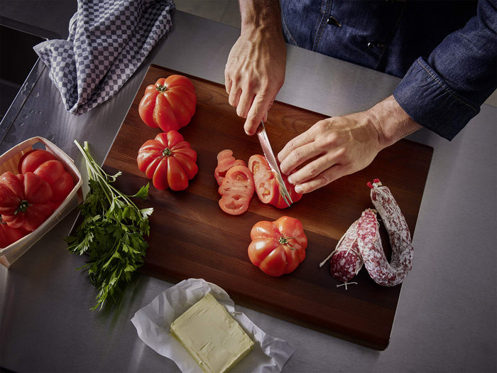 Individual cutting tomatoes on a cutting board next to herbs, butter, and sausage